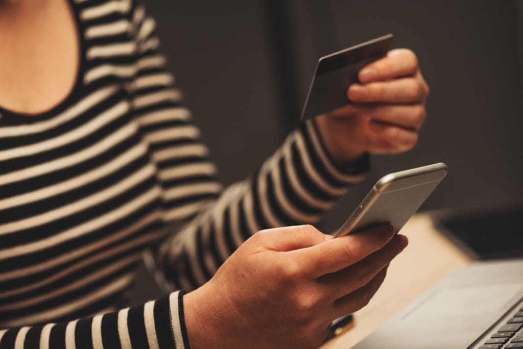 A imagem mostra uma mulher com o celular e um cartão nas mãos. Ela está utilizando o m-payment.