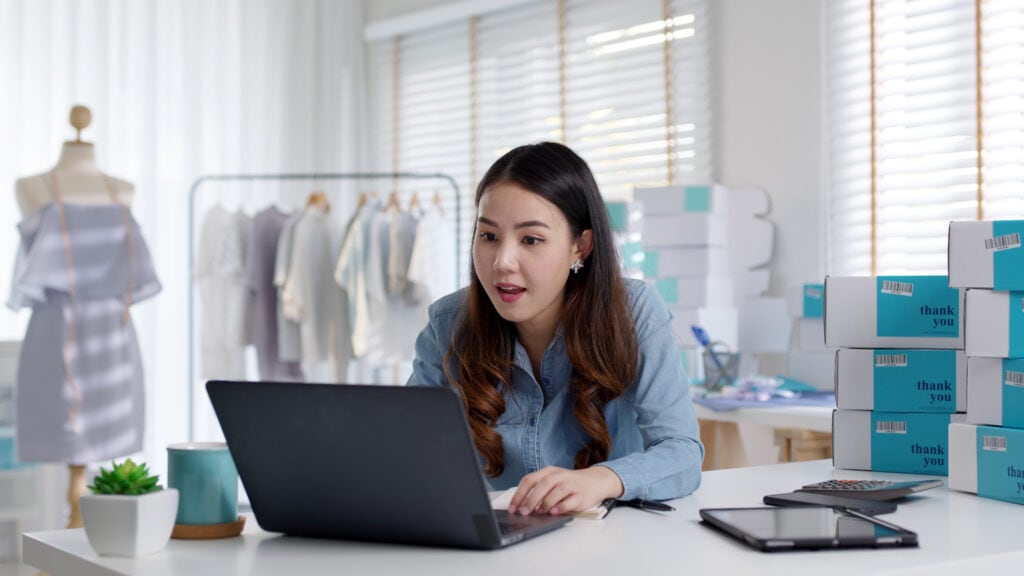 A foto mostra uma mulher, ela está sentada em frente a mesa. Na mesa tem um computador, celular, caixas de produtos vendidos. Ela está procurando Como gerar boleto para clientes.