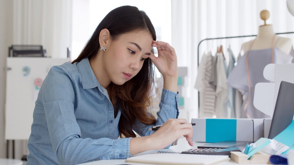 A imagem tem um mulher, ela está sentada em frente a uma mesa, na mesa tem papéis, caneta e uma calculadora. Ela está com a mão na testa e a outra digitando na calculadora, fazendo o fluxo de caixa de forma manual.