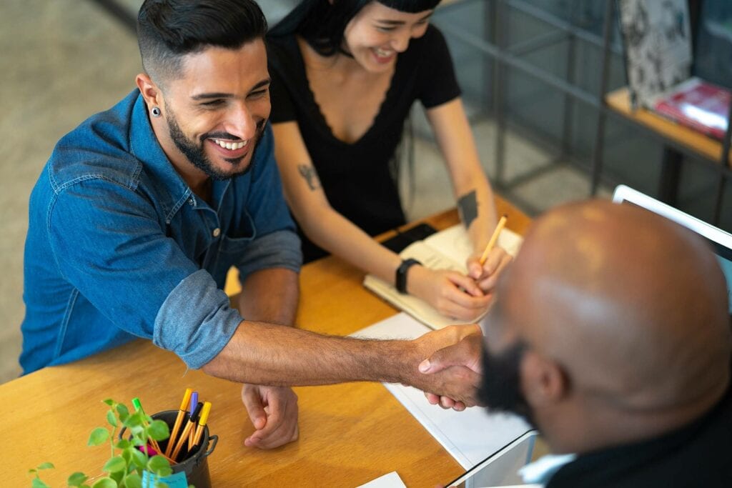 A satisfação do cliente está representada na imagem. No cenário há uma mesa com algumas folhas encima. Deum lado da mesa há um casal, o homem tem cabelos pretos e barba e está sorrindo, ele veste uma blusa jeans. A mulher, tem cabelos pretos e veste um vestido preto, ela está sorrindo. Ambos apertam as mãos de um homem que está do lado oposto da mesa. Ele é careca e usa óculos e veste um terno preto.