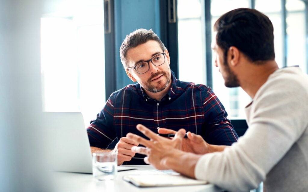 Dois homens sentados lado a lado em frente a uma mesa. Na mesa tem um copo de água, alguns papéis e um notebook. Um dos homens está usando óculos e uma blusa xadrez e o outro está usando uma blusa branca. A imagem busca representar uma conversa fazer cobrança de clientes.