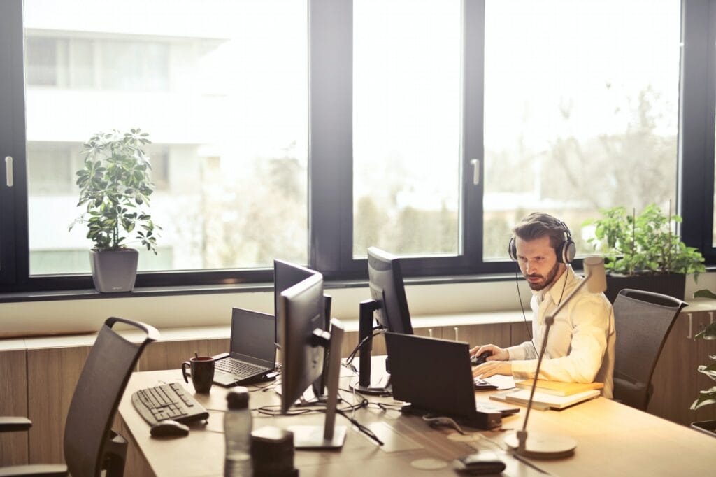 Homem está sentado em uma cadeira no escritório, fazendo atendimento ao cliente. Ele veste uma blusa branca e está com fones no ouvido. Em sua mesa há 2 notebooks e dois monitores, itens de informática e acessórios de escritório.