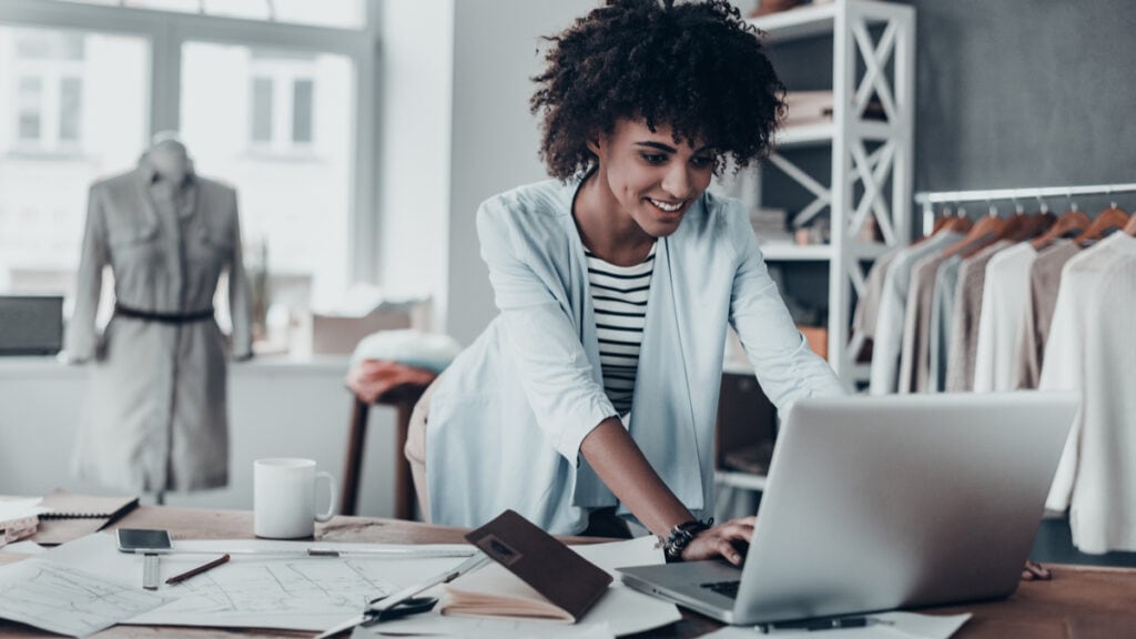 Uma mulher negra está em seu ateliê trabalhando. A mulher veste uma blusa listrada com um suéter cinza por cima. Em sua mesa, contém um notebook, papéis, caneca e outros materiais de escritório. Ao fundo, estantes e araras com roupas. Ela está feliz por utilizar um erp saas e gerenciar seu negócio.