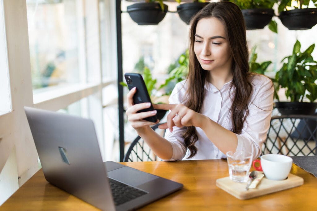 Na imagem, temos uma mulher em seu escritório, com um celular nas mãos e um computador sobre a mesa. A mulher usando a api de pagamento.