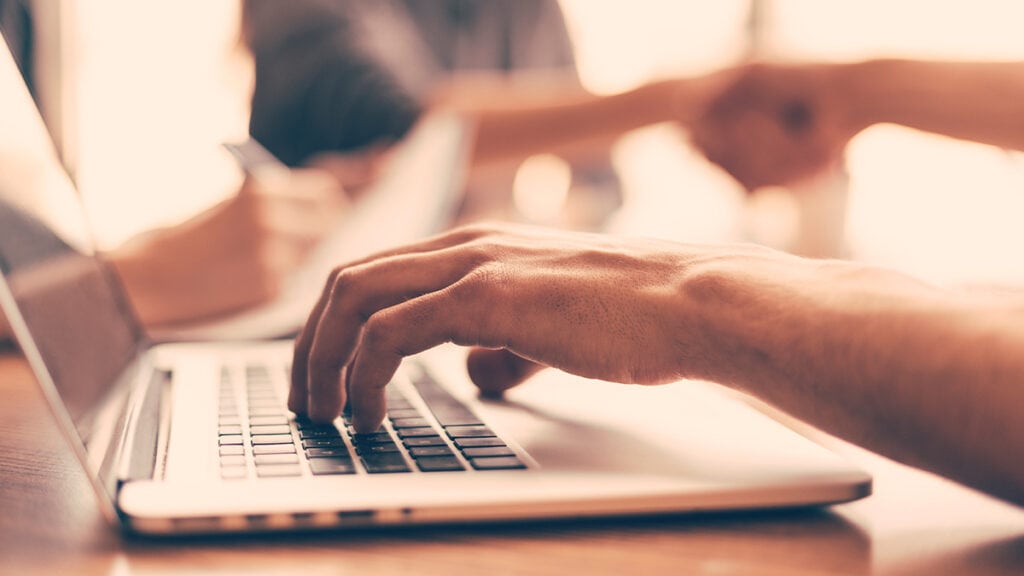 Na imagem, um mão masculina está digitando em um notebook. A imagem também possui tons pastéis e ao fundo aparecem flashs de outras pessoas presentes em um escritório. Ela representa o processo de cadastro de clientes.