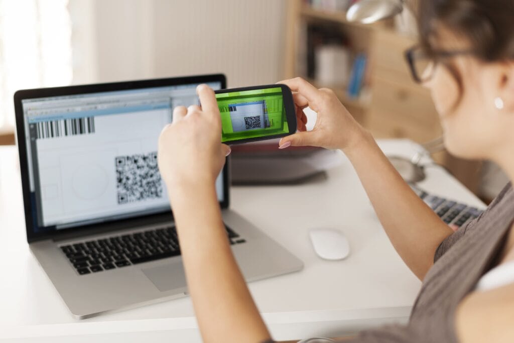 A imagem tem um homem, sentado em frente a uma mesa com um computador, ele segura um celular na mão e está fazendo um pix no e-commerce.