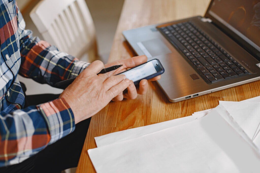 A imagem mostra as mãos de um empreendedor mexendo no celular, em frente ao computador. Ele está verificando o recebimento do boleto de cobrança.