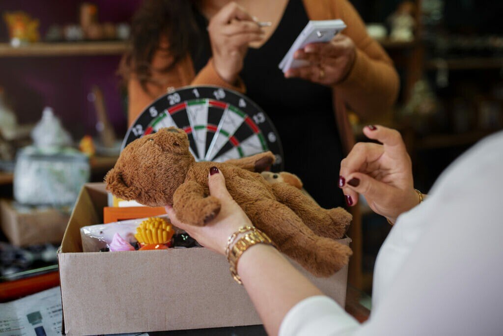 A imagem mostra duas vendedoras de uma loja, em volta de uma caixa com diversos brinquedos. Uma delas está segurando um bloco de notas e a outra um urso de pelúcia. Elas estão conversando sobre como vender mais no dia das crianças.