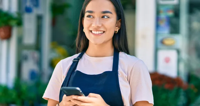 Mulher usa avental e está segurando um celular, representando comercial e vendas.